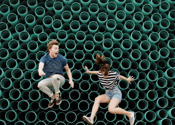 Two people jumping in front of a wall of green pipes